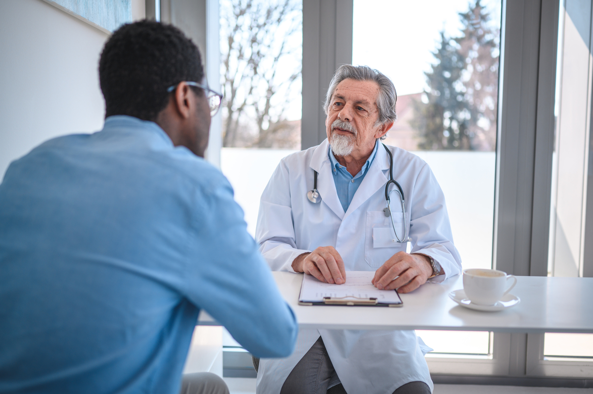  Waist up of a Black male patient in a meeting with a senior caucasian male doctor and ophthalmologist. They are talking and looking at each other at the doctor's office.