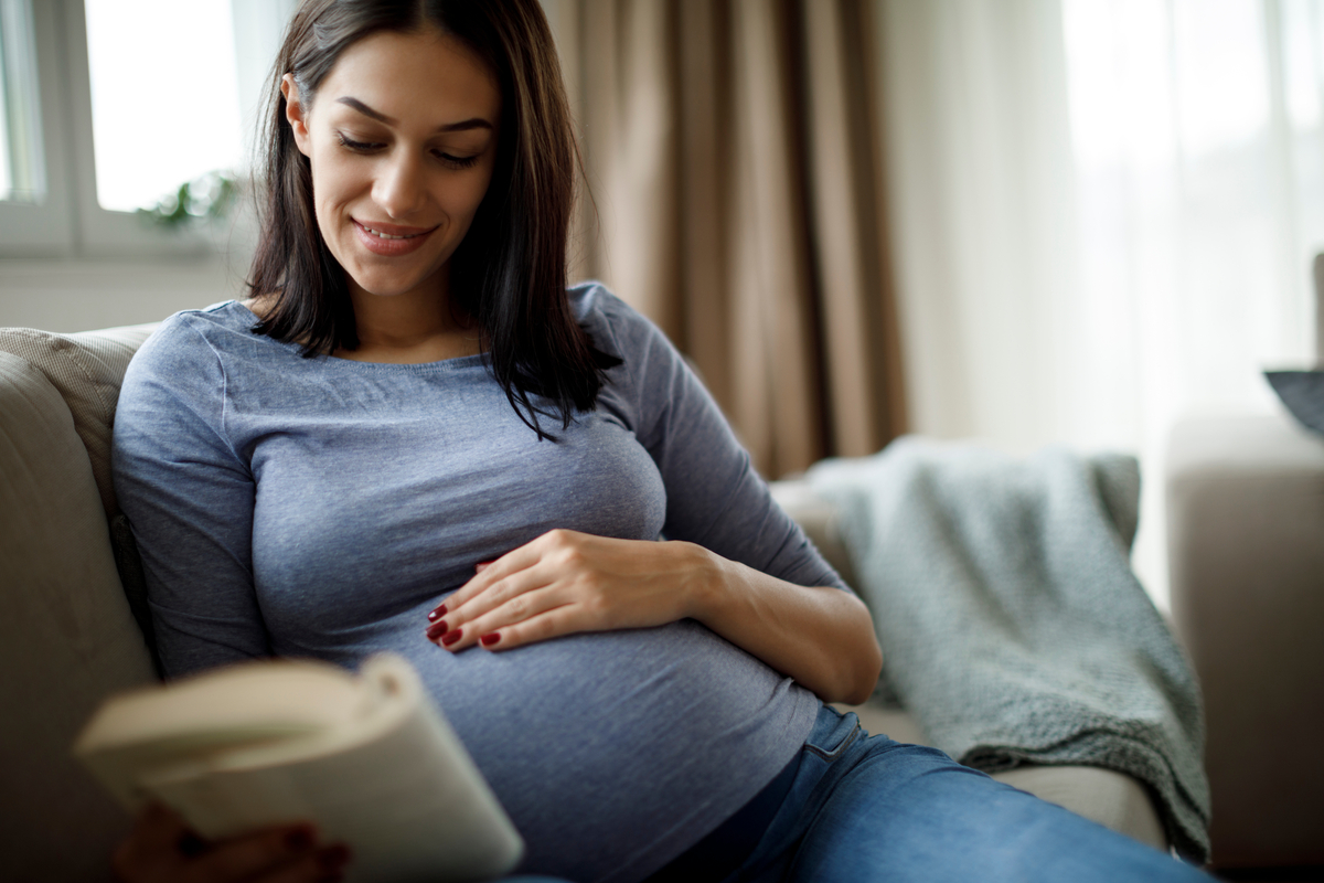 Pregnant woman sitting on a couch and she is looking at a book
