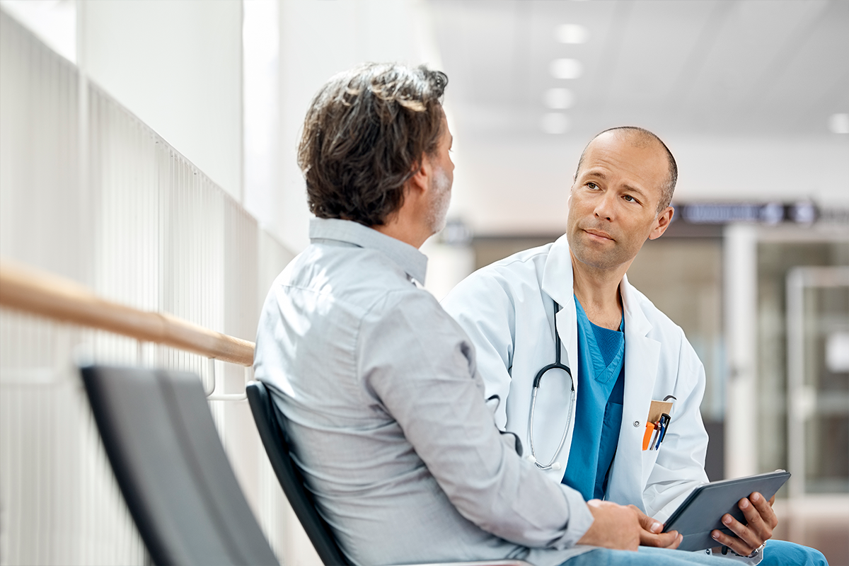 Male doctor counseling mature patient in waiting room. Medical professional is listening to ill man while holding digital tablet in hospital. They are sitting on seat.